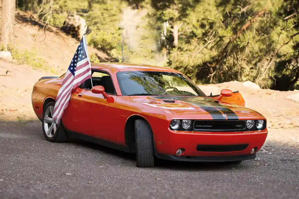 A red Dodge Challenger SRT with black racing stripes is parked on a gravel road surrounded by trees. An American flag is…
