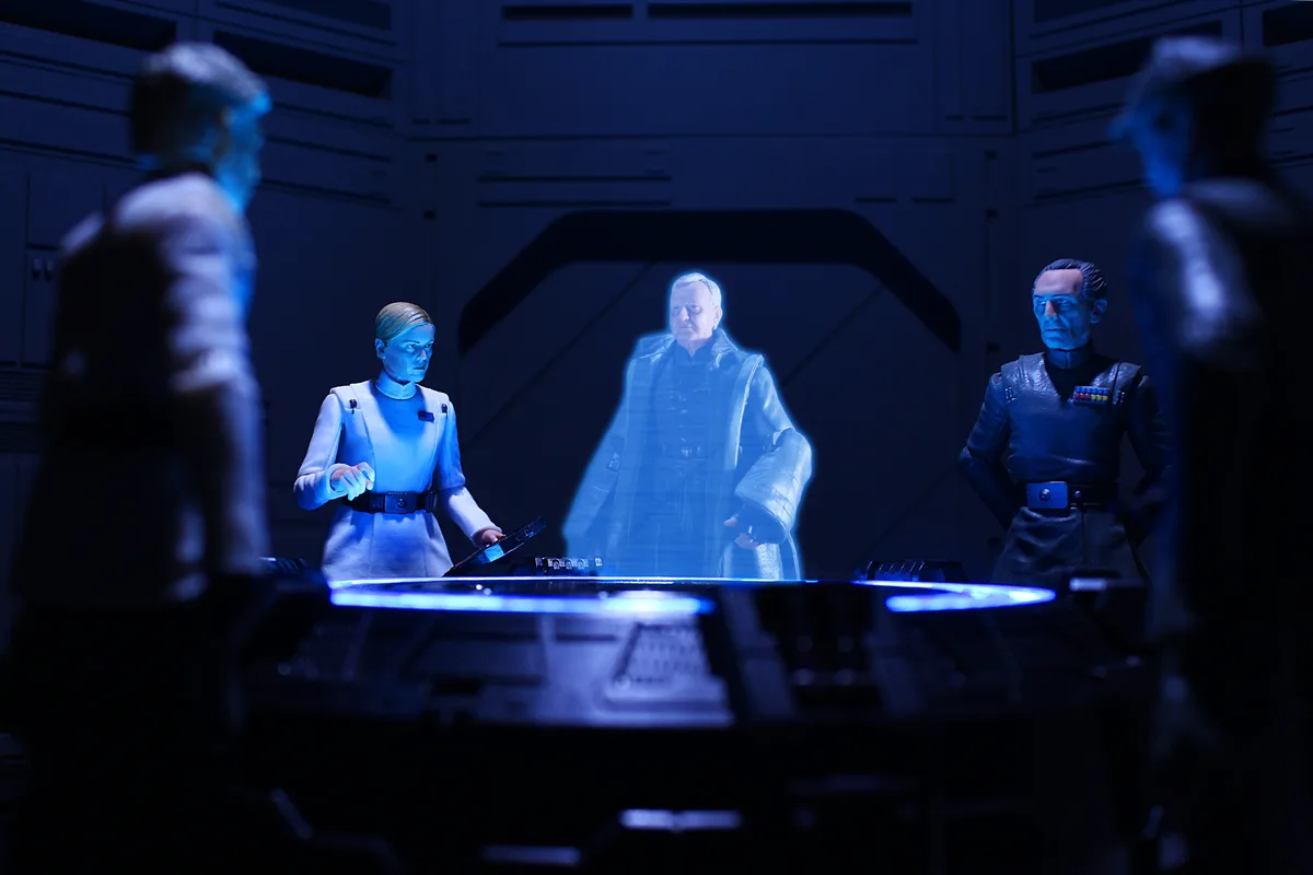 A still from a Star Wars scene featuring action figures of characters in a dimly lit, blue-lit room around a glowing table…