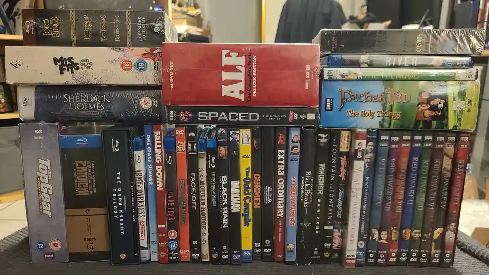 A large, cluttered stack of DVD and Blu-ray cases is arranged on a shelf, featuring titles like 'The Lord of the Rings'…