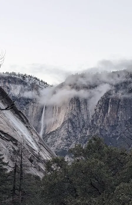 A misty mountain landscape with a waterfall cascading down a sheer cliff face, framed by trees in the foreground. The…