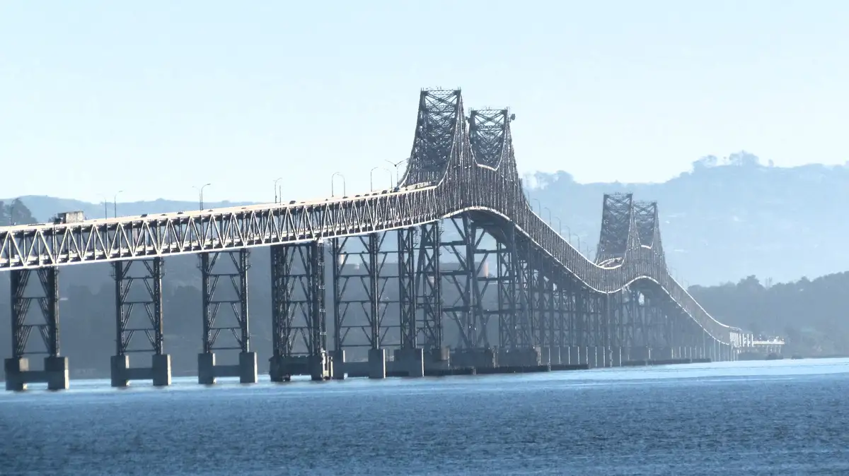 A wide shot of the Bay Bridge in San Francisco, California, stretching across a body of water with hills in the hazy…