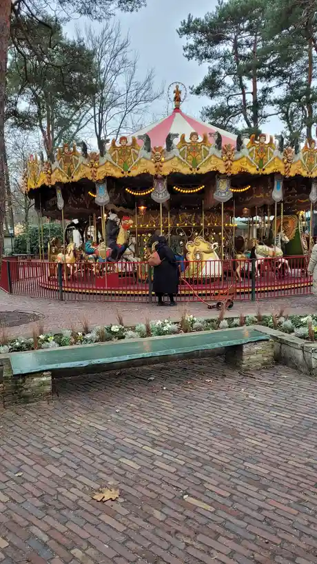 A person in a black coat stands outside a colorful, ornate carousel with a red fence, looking at it while a child rides a…