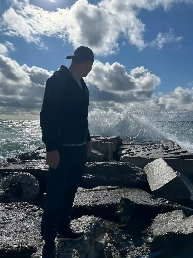 A person in a dark hoodie and cap stands on wet concrete blocks at a rocky shoreline, looking out at the ocean where waves…