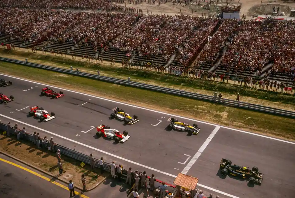 An aerial photo of a Formula 1 race with multiple cars on the track and a large crowd in the stands. The overlaid text reads…