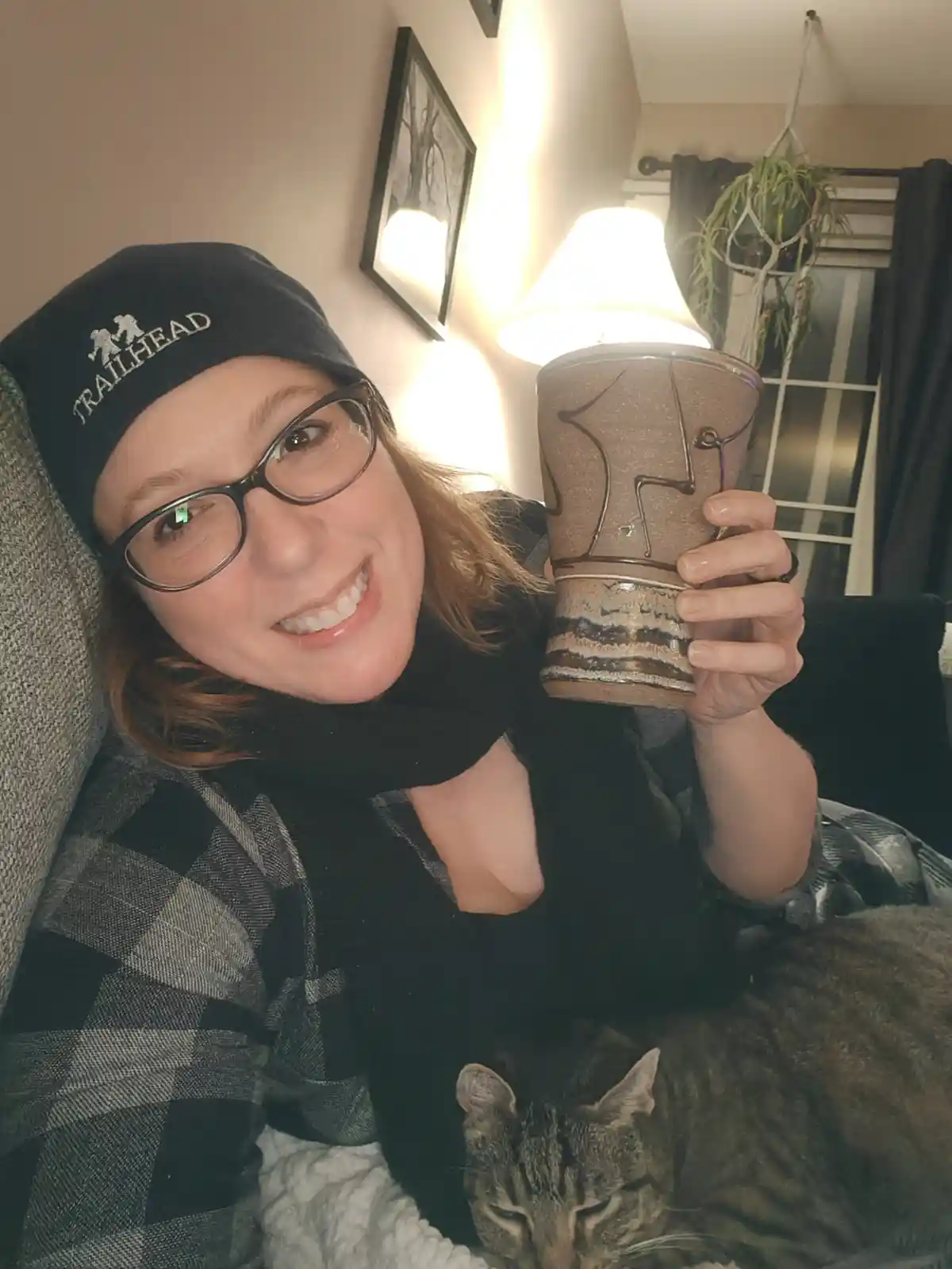 A smiling woman wearing glasses and a black 'Trailhead' beanie sits on a couch holding a large ceramic mug, with a sleeping…