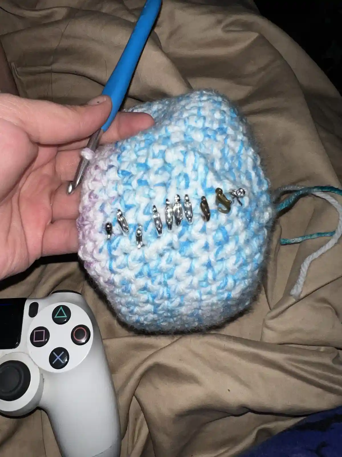 A hand holds a blue and white crocheted hat with a blue crochet hook inserted, and several safety pins are stuck into the…