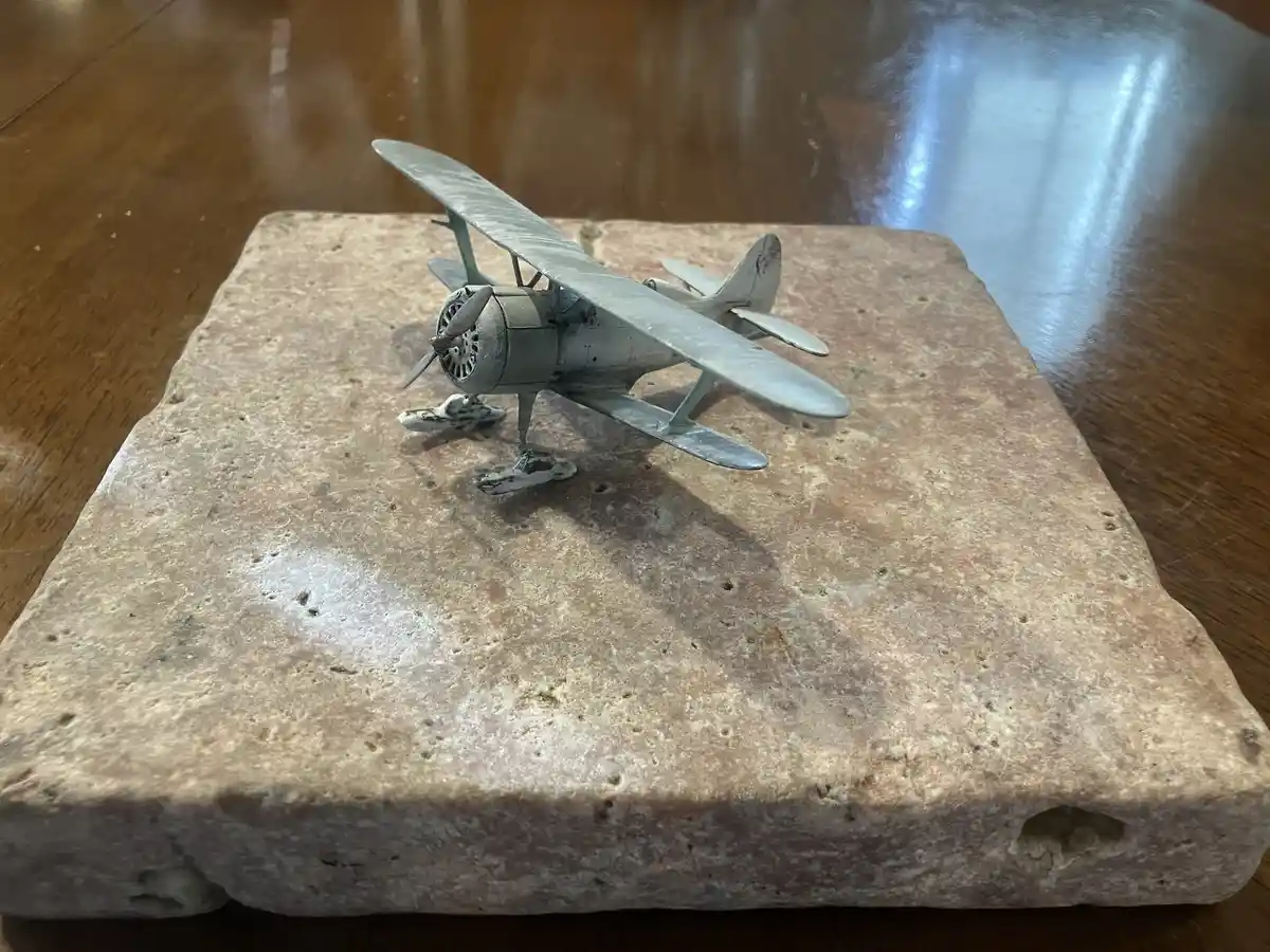 A small, weathered metal biplane model sits atop a rough, porous stone block resting on a wooden table. The overlaid text…
