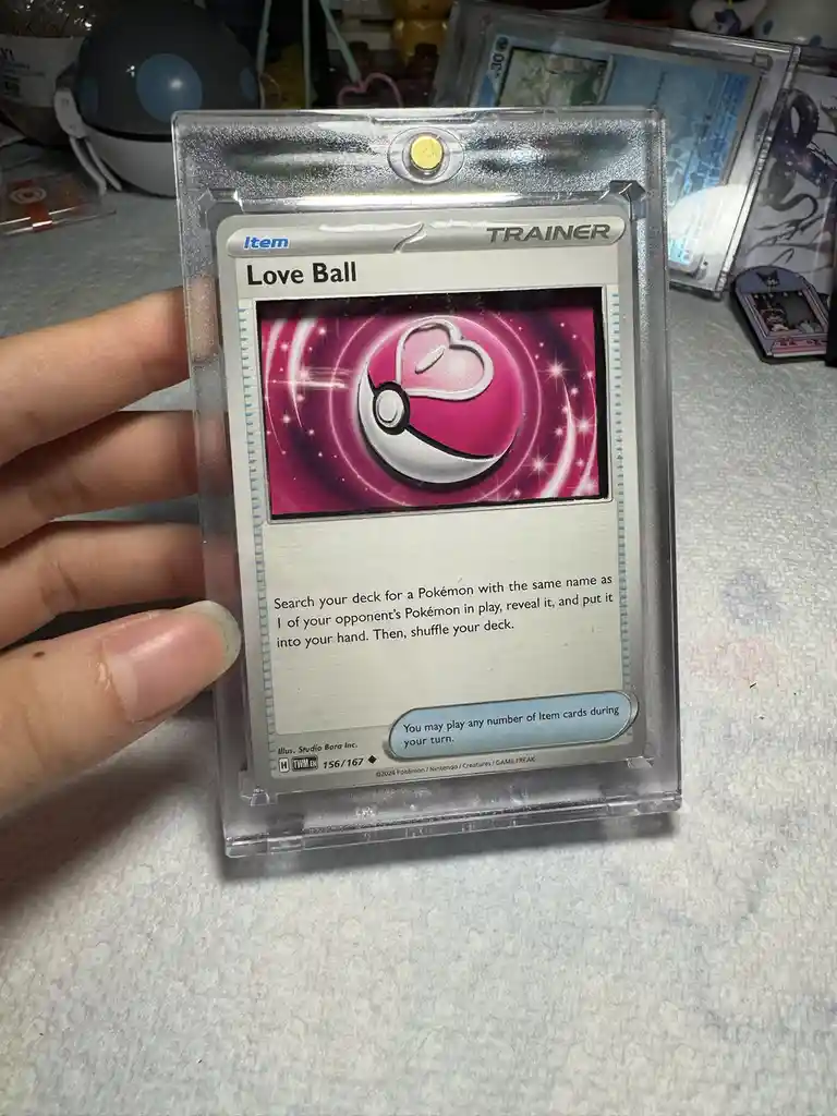 A hand holds a Pokémon TCG 'Love Ball' Item Trainer card in a clear protective sleeve. The card's artwork shows a pink Poké…