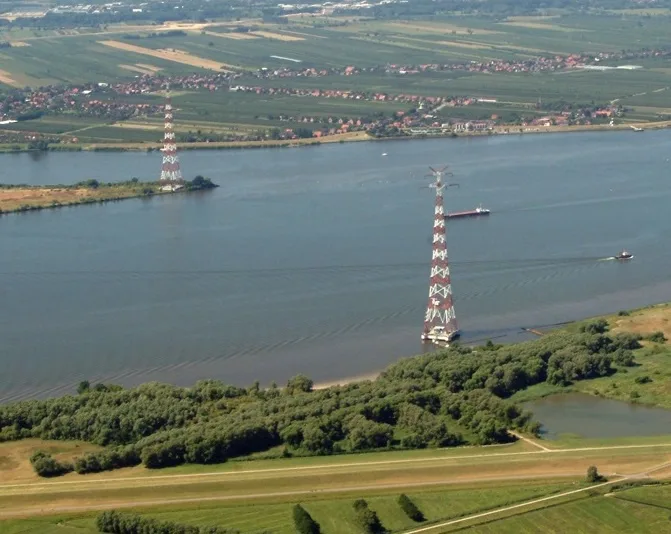 An aerial view shows two tall, red-and-white striped lattice towers standing in a wide river, surrounded by green fields and…