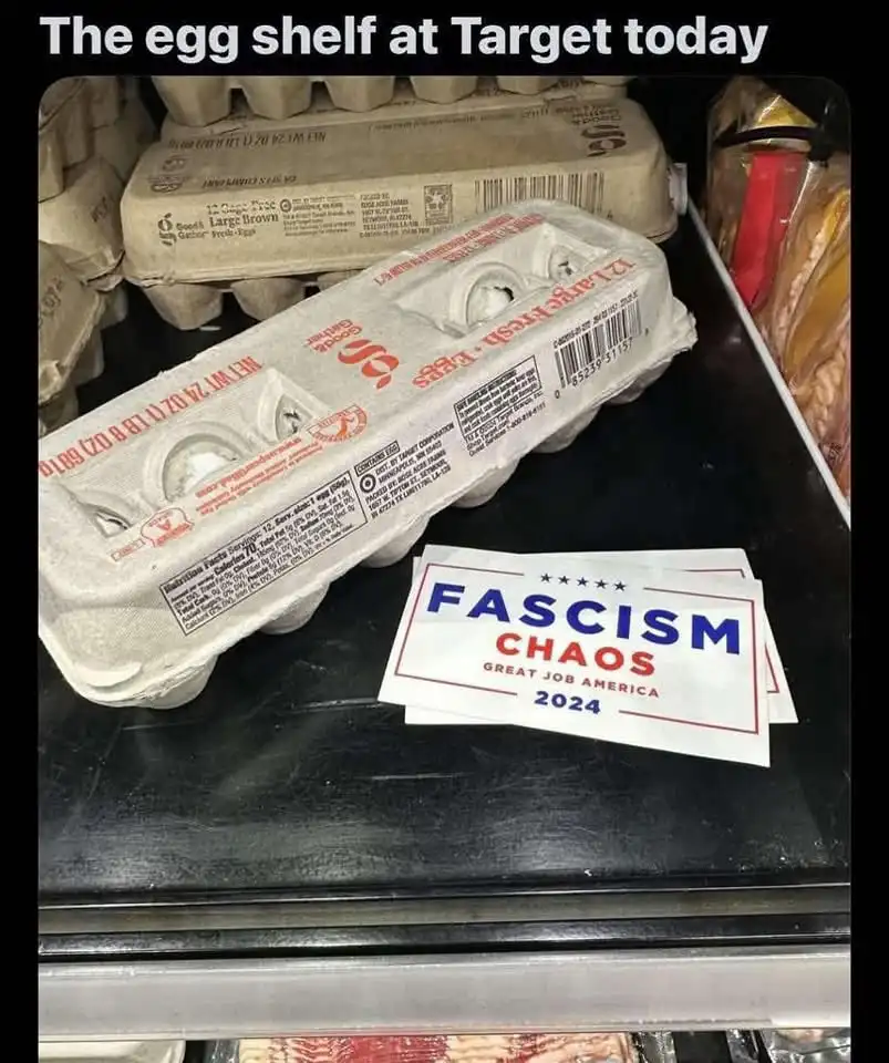 A photo of an egg carton on a supermarket shelf with a political-style sticker that reads 'FASCISM CHAOS GREAT JOB AMERICA…
