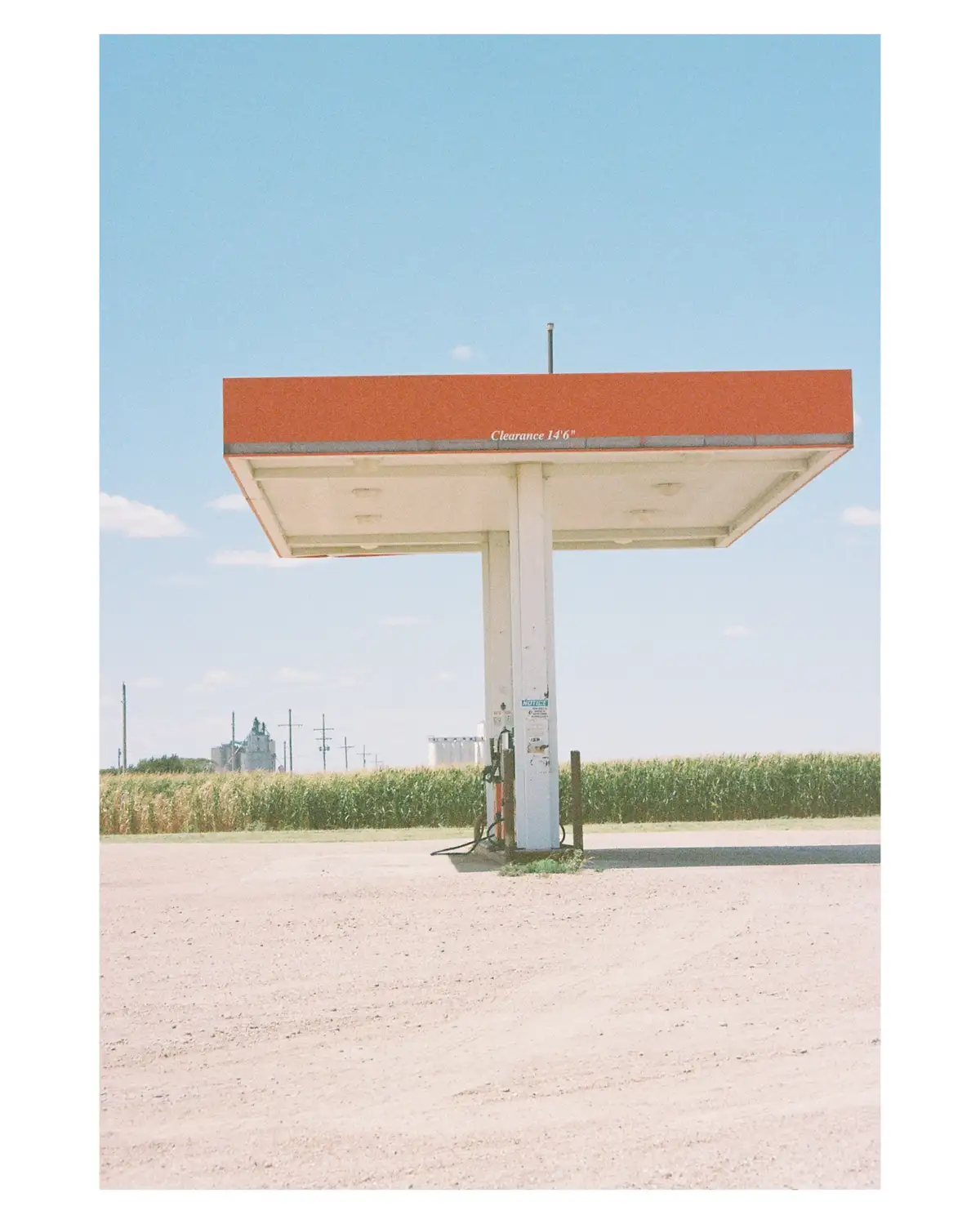 A lone, weathered gas station canopy stands in a vast, empty field under a clear blue sky, with a cornfield and distant farm…