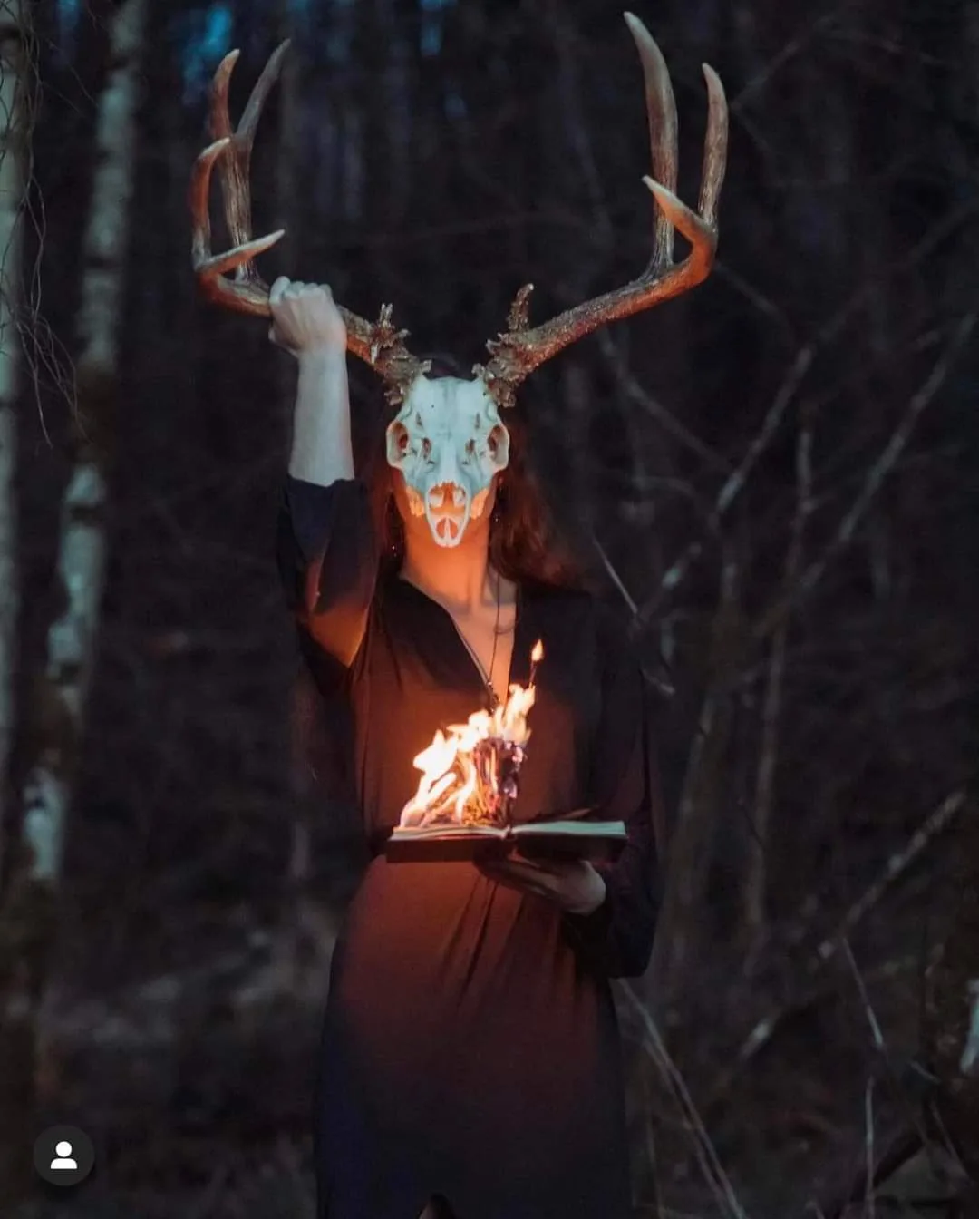 A person in a dark forest wears a deer skull mask with antlers, holding up the antlers with one hand while holding an open…