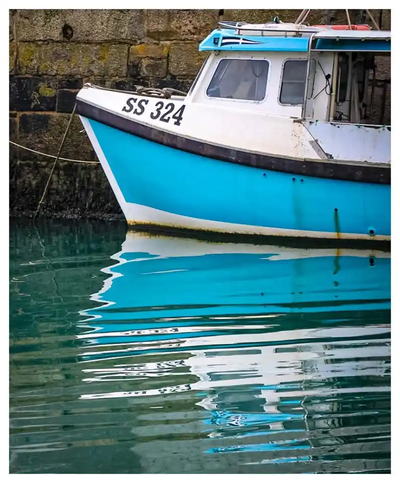 A blue and white fishing boat with the hull number 'SS 324' is moored next to a stone wall, its reflection rippling in the…