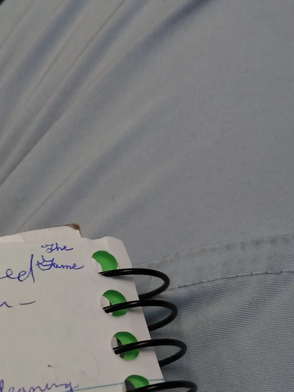 A close-up of a spiral-bound notebook resting on a light blue fabric surface, showing handwritten text that reads 'The Game'…