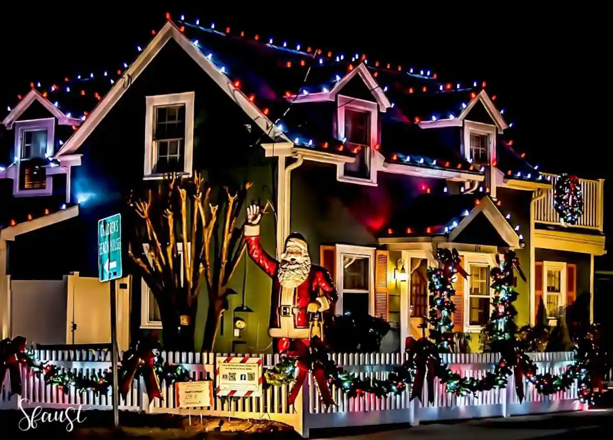 A house at night is extravagantly decorated with colorful string lights along the roofline and festive garlands on a white…