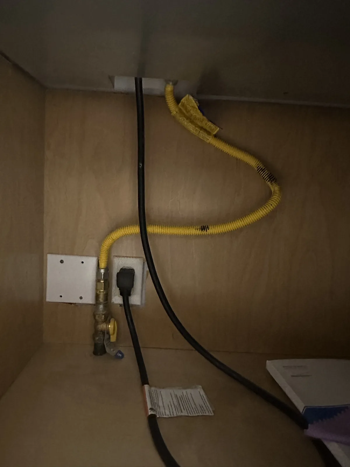 A dimly lit under-cabinet view shows a yellow flexible gas hose connected to a brass valve and an electrical outlet, with a…