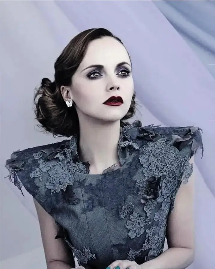 A woman with dark, dramatic eye makeup and red lipstick stares intensely at the camera while wearing a dark, ornate…