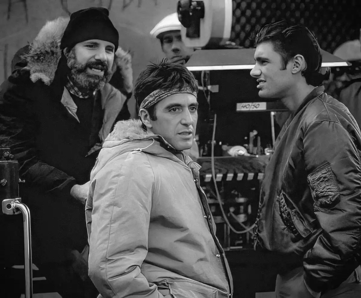 A black-and-white photo from a film set shows three men. On the left, a bearded man in a winter coat and beanie smiles. In…