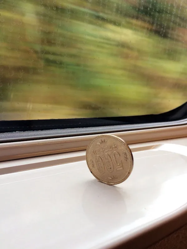 A gold-colored 100 yen coin stands upright on a white train window sill, with a blurred green landscape visible through the…