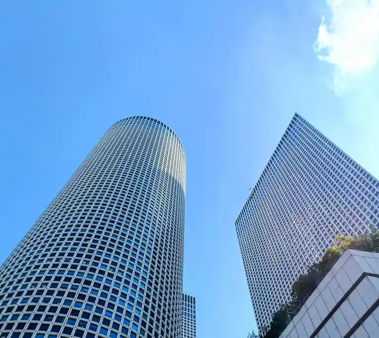 A low-angle photo shows two modern skyscrapers against a clear blue sky with a few clouds. One building is cylindrical and…