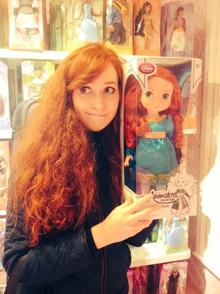 A woman with long, wavy red hair stands beside a Disney Store box containing a Merida doll, both sharing identical red hair…