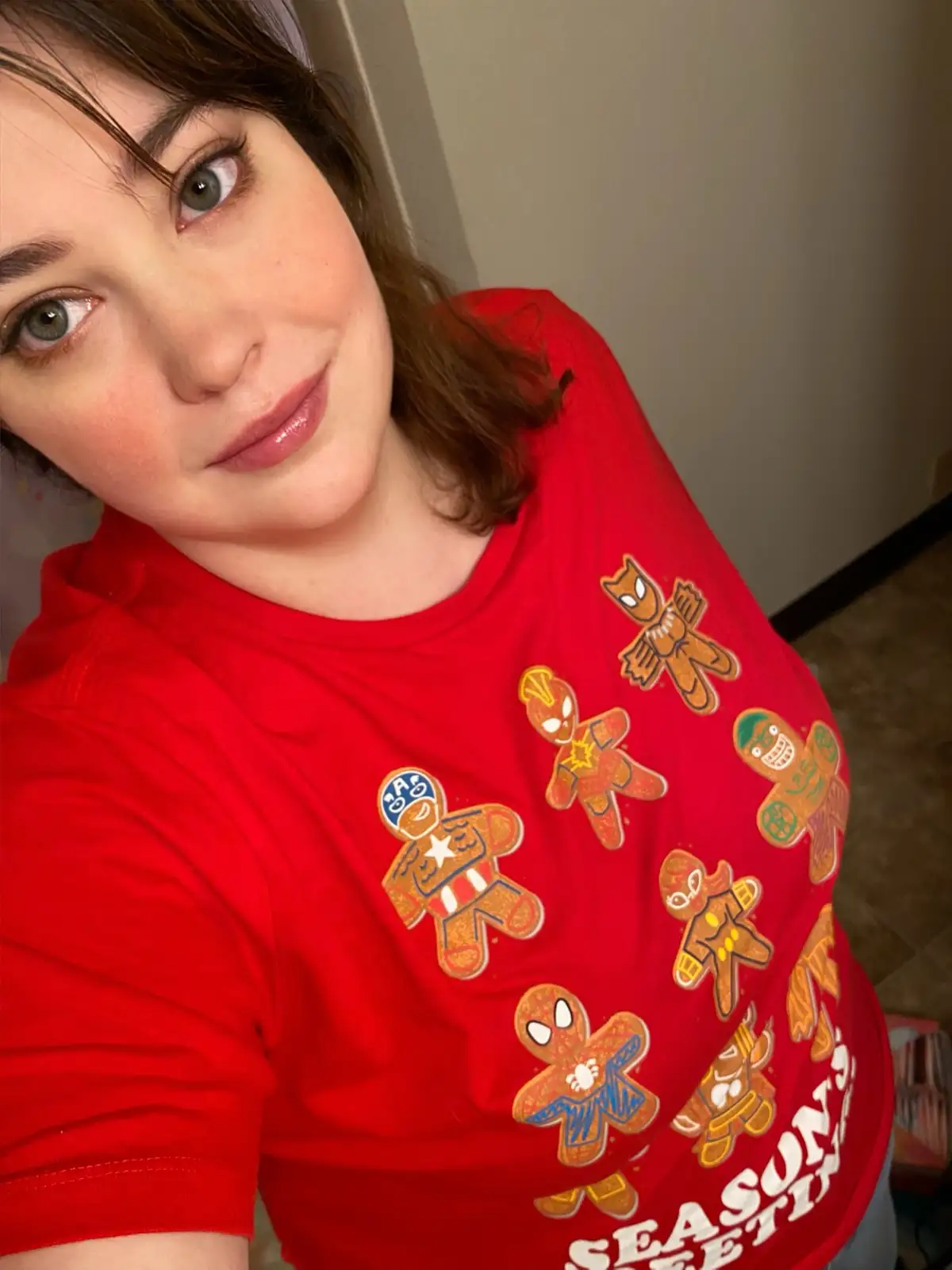 A person with brown hair and green eyes looks at the camera while wearing a red t-shirt covered in gingerbread men decorated…