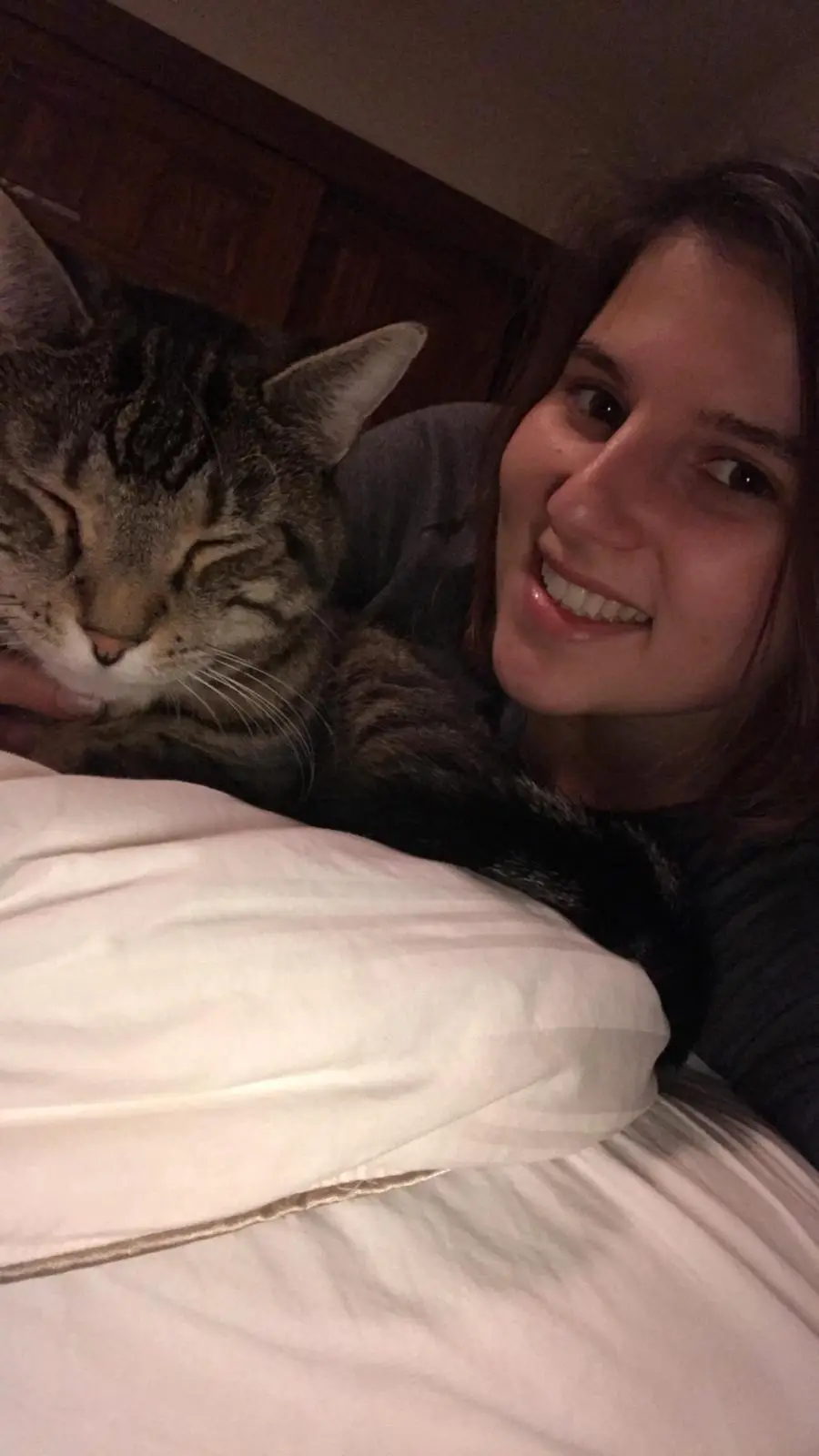 A person smiles at the camera while lying in bed with a tabby cat sleeping peacefully on their chest. The cat has its eyes…