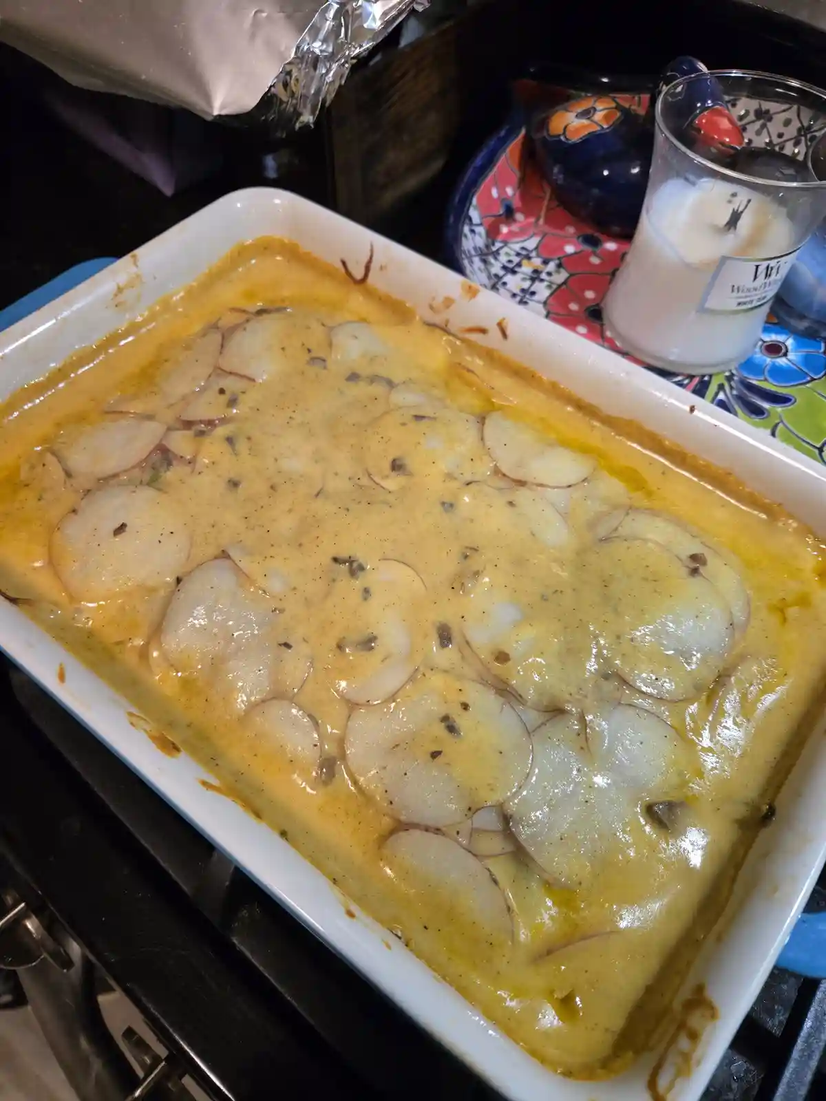 A white rectangular baking dish filled with a creamy yellow sauce topped with thinly sliced potatoes, resting on a stovetop…