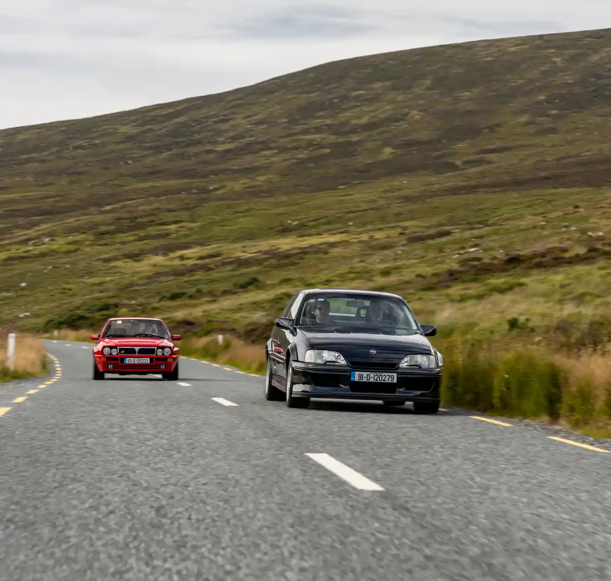 Two cars drive on a winding rural road through hilly, grassy terrain under an overcast sky. The car in the foreground is a…