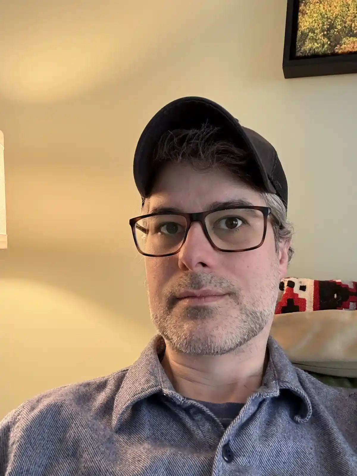 A man with glasses and a black baseball cap looks directly at the camera with a neutral expression, wearing a blue button-up…
