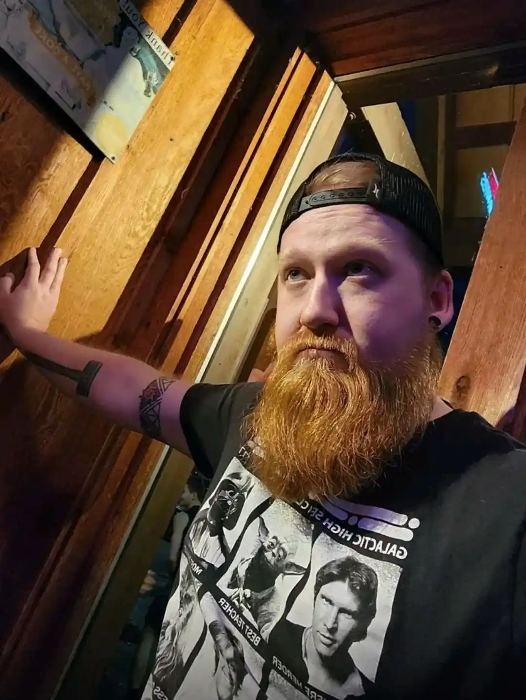 A man with a long red beard and black cap wears a black t-shirt featuring Star Wars characters like Yoda, Darth Vader, and…
