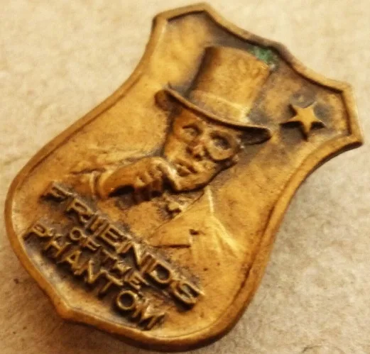 A close-up of a bronze-colored, shield-shaped pin featuring a raised relief of a man in a top hat and glasses, with a star…