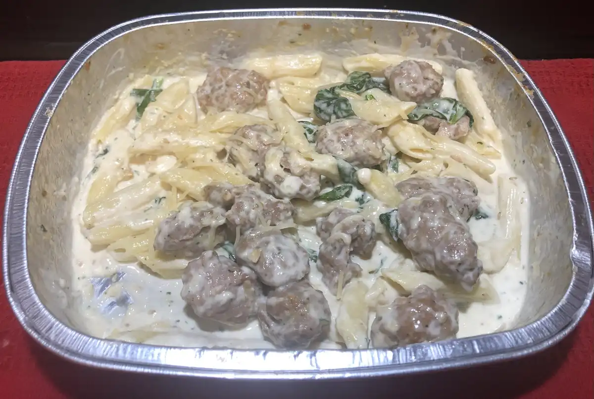 A close-up photo shows a rectangular aluminum baking pan filled with creamy pasta, penne noodles, spinach, and numerous…