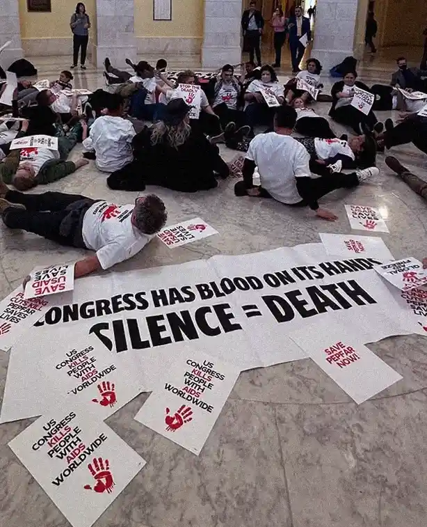A group of protesters lie on the marble floor of a government building, holding signs that say 'SAVE US AIDS' and 'US…