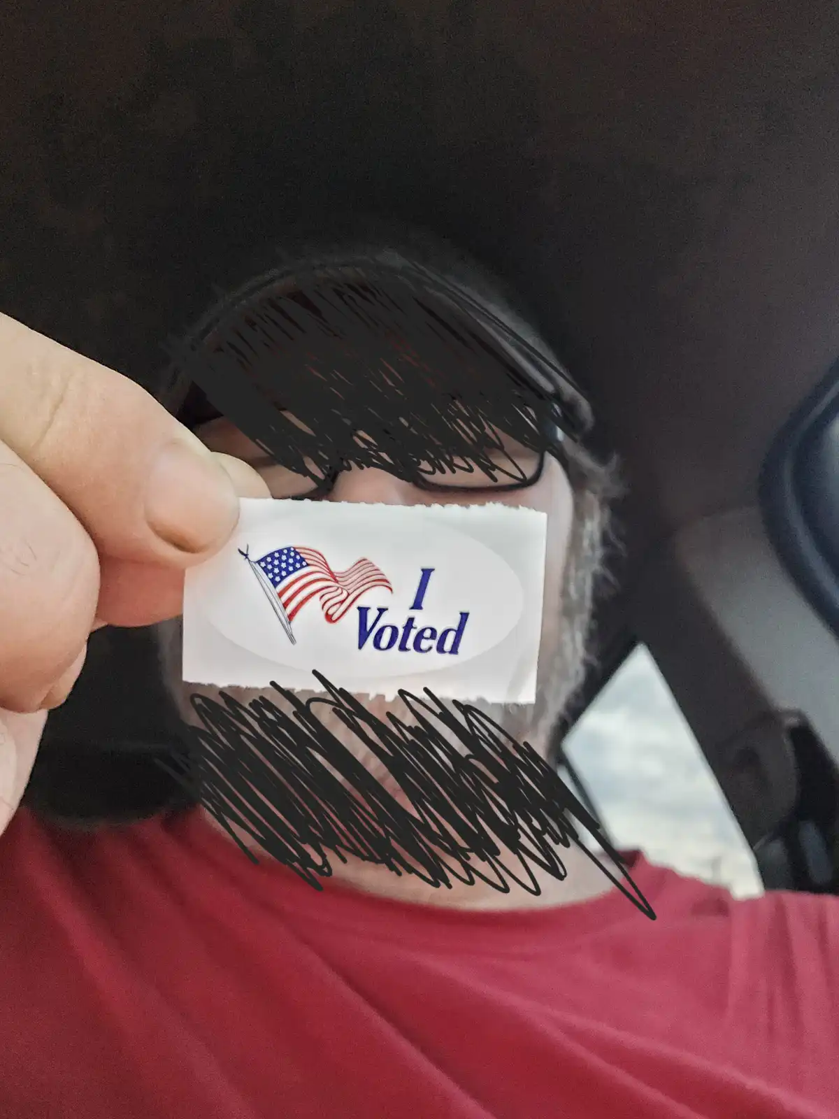 A person wearing glasses and a red shirt holds up an 'I Voted' sticker, obscuring their face with black scribbles over their…