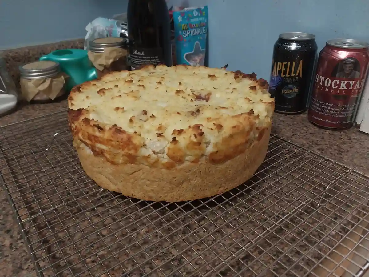 A round, golden-brown baked dish sits on a metal cooling rack on a granite countertop, surrounded by beer cans, a wine…