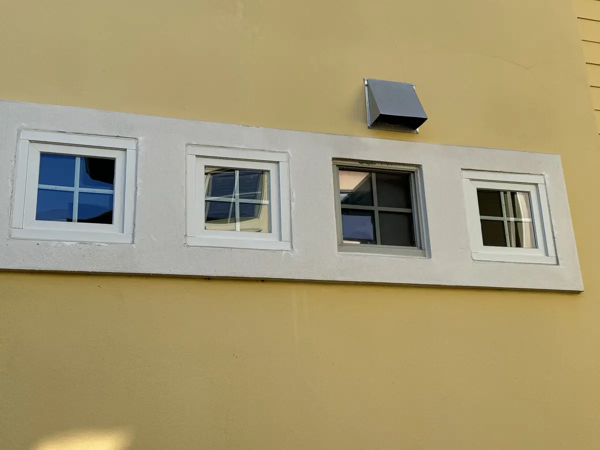 A photo of four windows set in a white sill on a yellow building wall, with a metal vent above them. The text overlay reads…