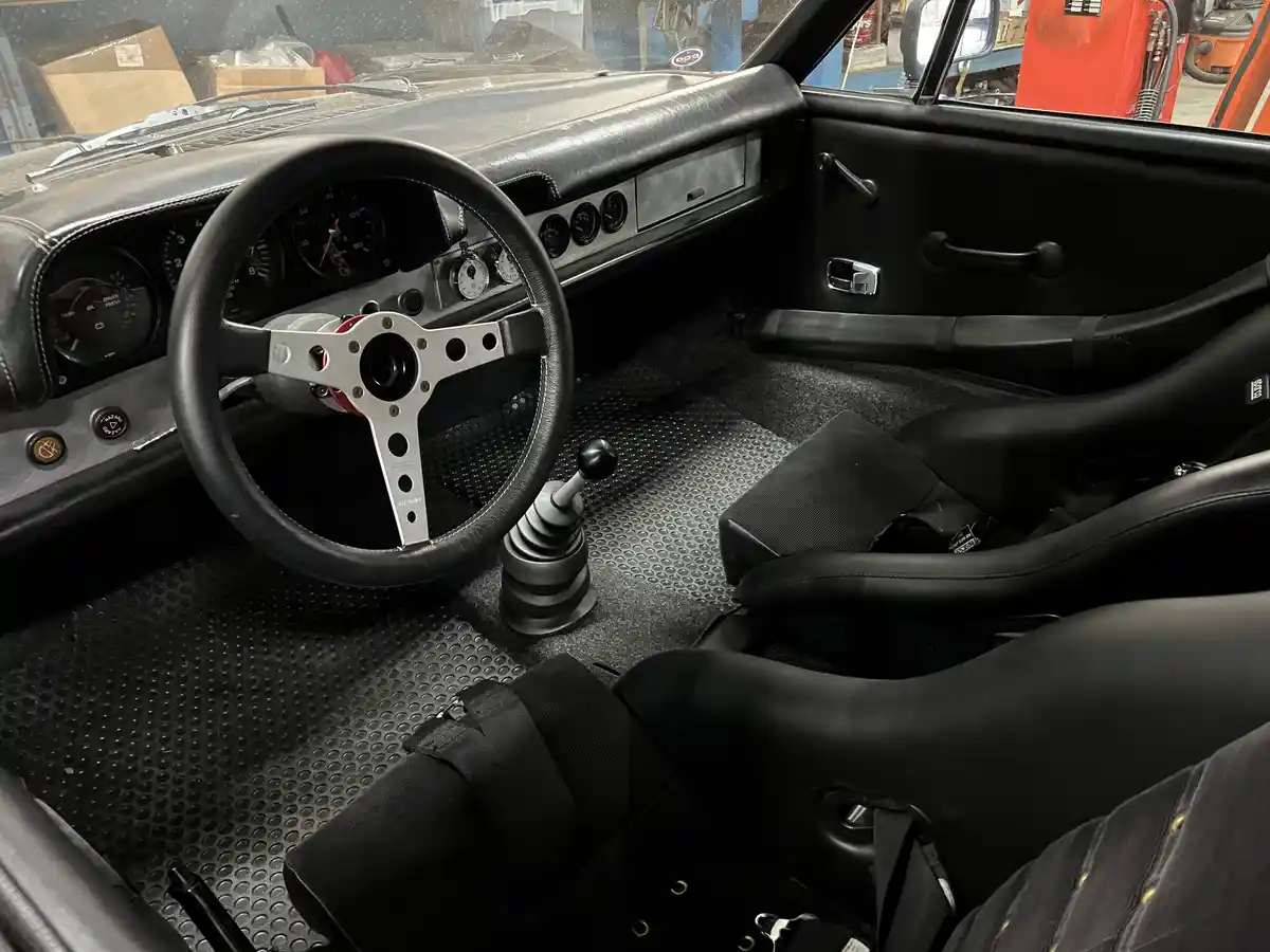 The image shows the interior of a classic race-prepped car with black leather seats, a three-spoke steering wheel, a manual…