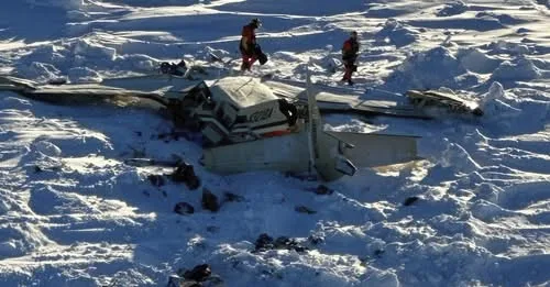 An aerial view shows the wreckage of a small white airplane buried in deep snow, with two rescuers in red gear walking…
