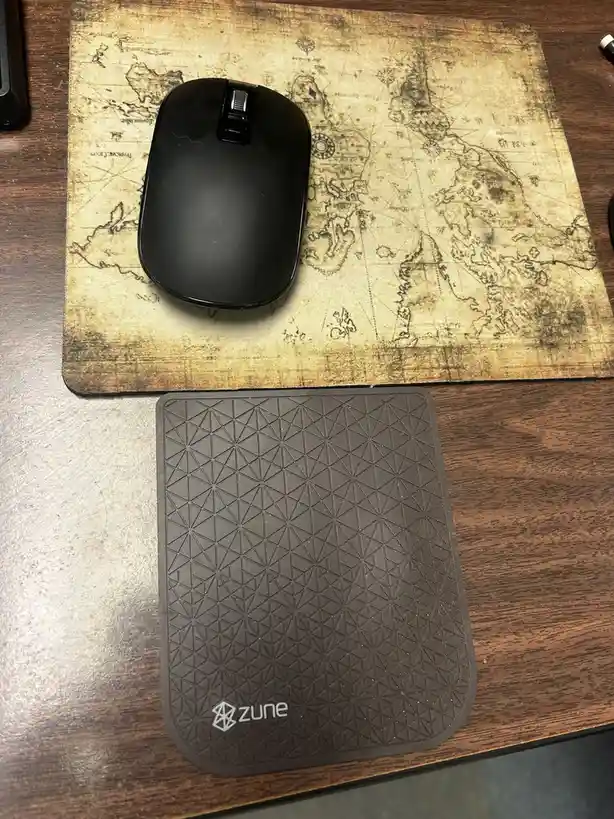 A black computer mouse sits on a vintage-style world map mousepad, positioned above a dark gray Zune-branded wrist rest with…