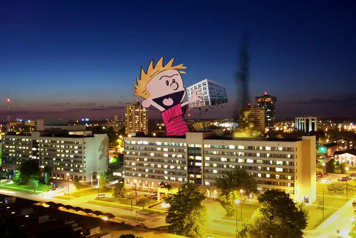 A nighttime cityscape photo of illuminated buildings shows a giant cartoon boy with spiky blonde hair and a red shirt…