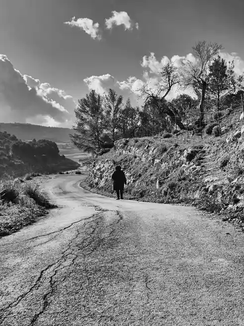 A black-and-white photo shows a person walking away down a cracked, winding rural road flanked by rocky hills and sparse…