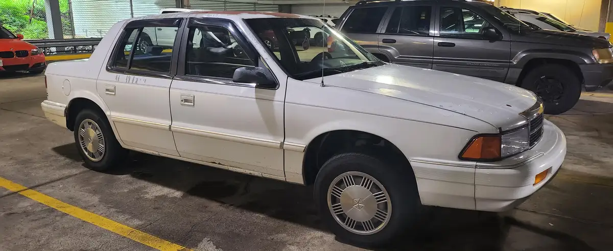 A white, visibly aged 1990s Mitsubishi sedan is parked in an indoor lot, showing rust, dirt, and wear. The overlaid text…
