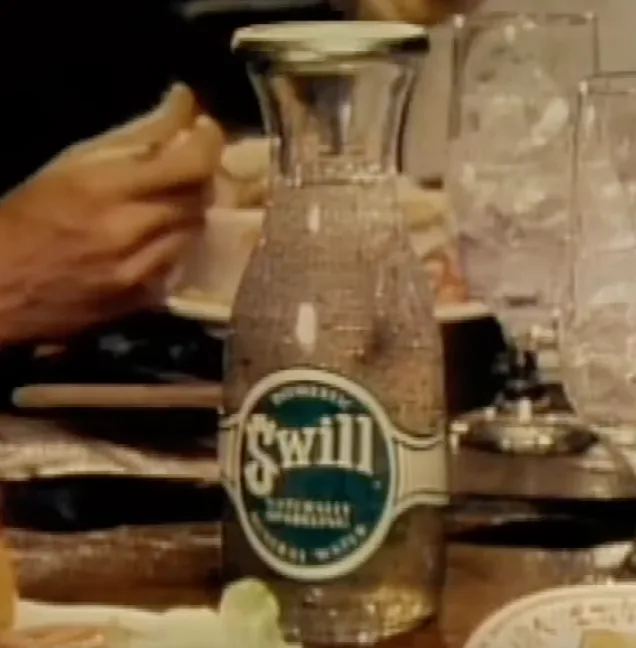 A vintage-style glass bottle labeled 'Swill Naturally Flavored Water' sits on a dining table, surrounded by glasses and a…