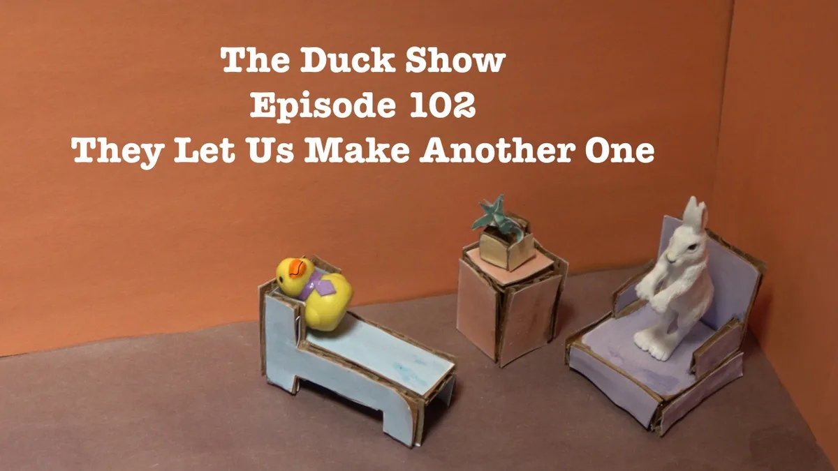 A miniature diorama shows a yellow rubber duck wearing a purple tie, reclining on a cardboard chaise lounge, while a white…