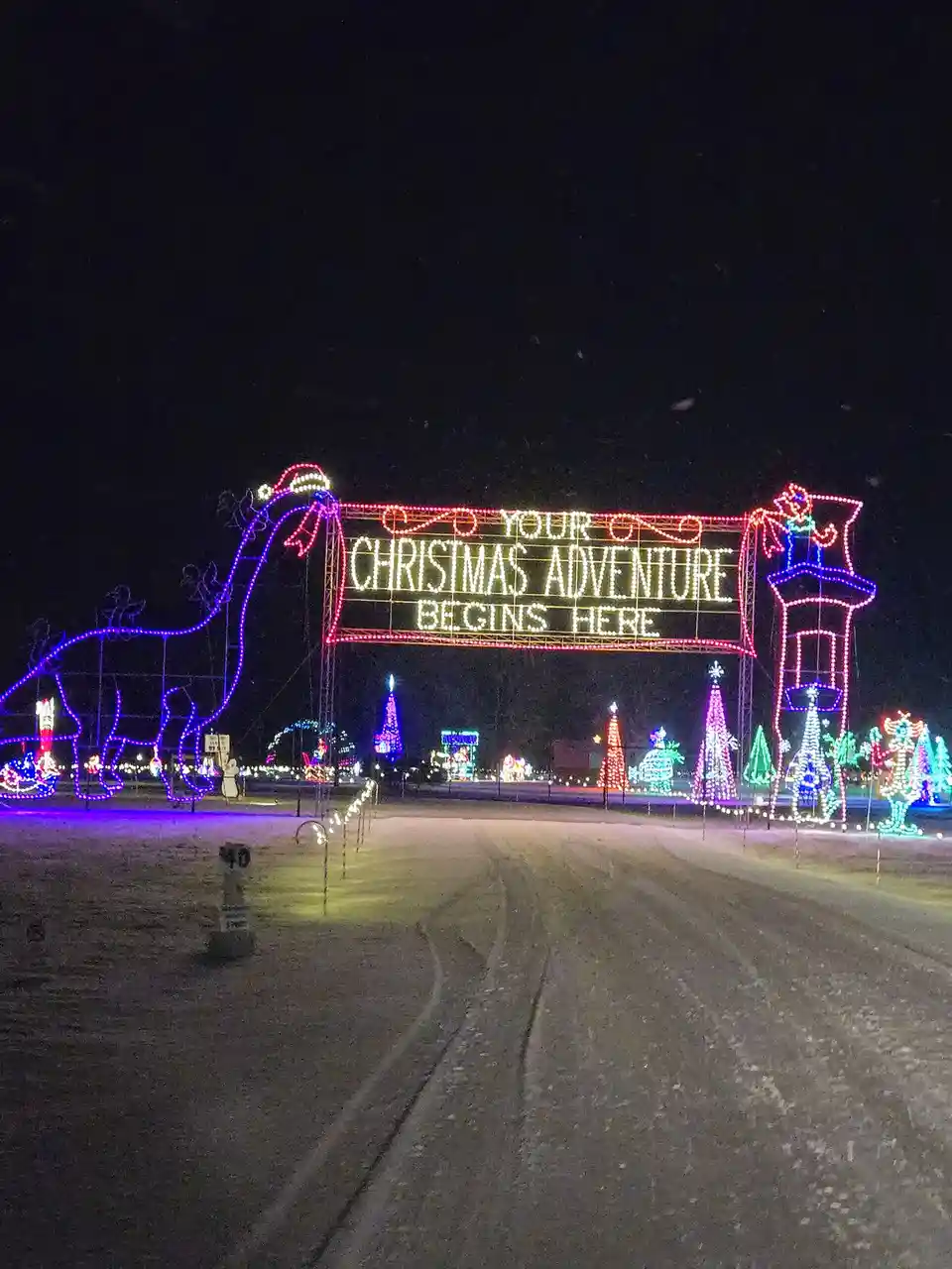 A nighttime photo of a large, brightly lit Christmas light display entrance with the text 'YOUR CHRISTMAS ADVENTURE BEGINS…