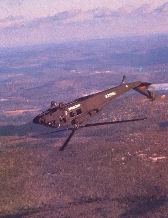 A vintage military helicopter, likely a UH-1 Huey, is shown flying over a hilly, forested landscape under a cloudy sky. The…