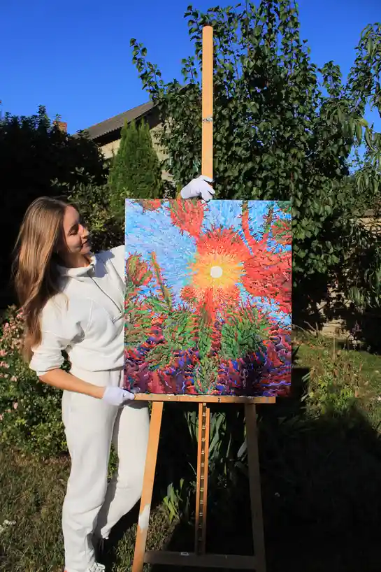 A woman in a white sweatsuit and white gloves stands outdoors beside an easel, holding up a vibrant, abstract painting of a…