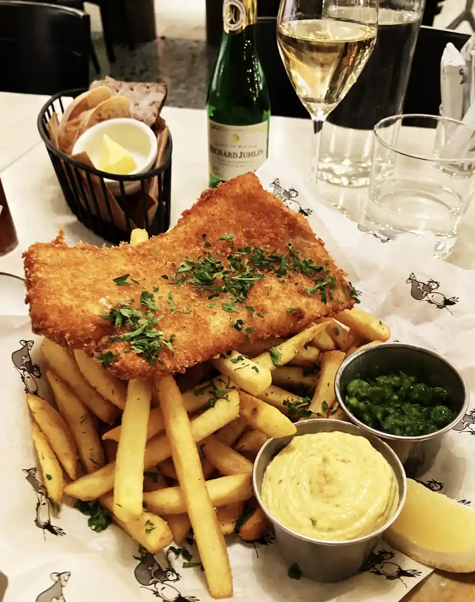 A close-up photo of a plated fish and chips meal featuring a large, golden-brown battered fish fillet resting atop a…