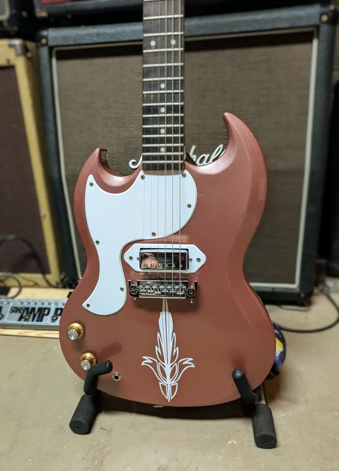 A reddish-brown electric guitar with a white pickguard and a white corn cob decal on its body stands upright on a stand…